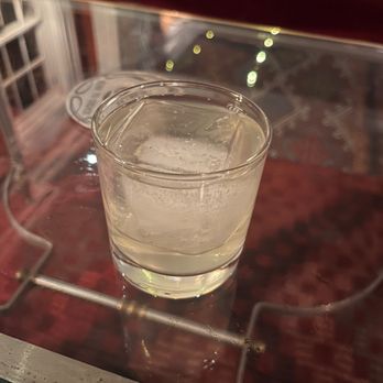 Argos Inn - clarified lassi - A transparent glass containing clarified lassi with ice cubes is positioned on a glass display case surface. The background is blurred, showing reflections and a patterned rug, indicating an indoor setting, possibly a restaurant or bar. - Hotels Near Me - Ithaca, New York