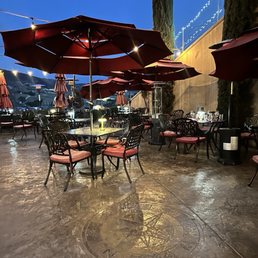 The image shows an outdoor dining area with multiple tables and chairs, each shaded by large red umbrellas. The ground is wet, reflecting the ambient lighting, and a compass rose is embedded in the concrete floor. String lights are hung overhead, and tall trees are visible in the background. The setting appears to be at dusk or early evening, with a clear sky and some distant landscape features visible.