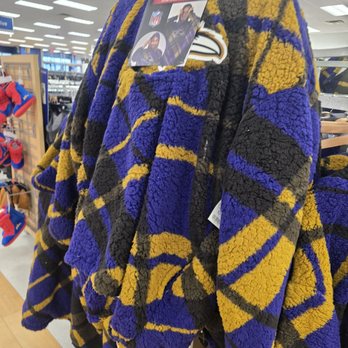 Marshalls - Go Ravens! - A fleece jacket with a multicolored abstract pattern in purple, yellow, and black is hanging on a display rack at Marshalls. The jacket has a tag attached, indicating it is a wearable NFL item. The background shows store lighting and other merchandise, including shoes, in a retail environment. - Department Stores Near Me - Gambrills, Maryland