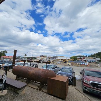Mad Jack's Mountaintop Barbecue - Beautiful Weather - A parking lot scene with several parked cars, an old rusted tank, and a partly cloudy sky overhead. - Barbeque Near Me - Cloudcroft, New Mexico