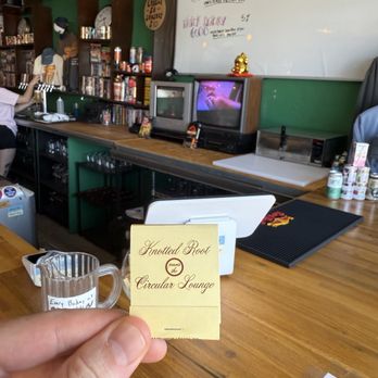 Circular Lounge - Cool matchbooks! - A person holds a business card for Knotted Root and the Circular Lounge inside a cafe or lounge. The background features a wooden counter with various items, including a small pitcher, a whiteboard with handwritten notes, and a vintage television. The setting appears casual with shelves filled with books and decorative items, indicating a relaxed atmosphere. - Lounges Near Me - Denver, Colorado