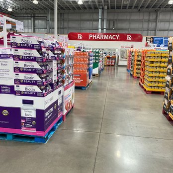 Costco - The image shows an aisle inside a Costco warehouse store, with shelves on both sides filled with various products, including health and beauty items. The store has a high ceiling with industrial lighting, and a prominent red and white sign indicating the pharmacy area at the back of the aisle. The floor is made of polished concrete, and the overall environment appears spacious and organized. - Wholesale Stores Near Me - Chicago, Illinois