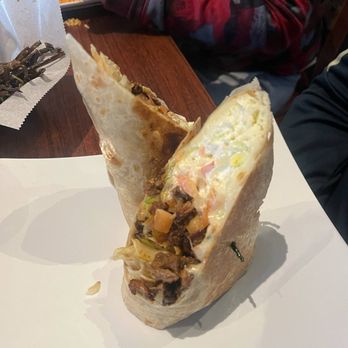 Taco City - Burrito - A burrito is cut in half and placed on a white sheet of paper, revealing ingredients like meat, vegetables, and sauce inside. The burrito has a soft flour tortilla exterior. In the background, a person wearing a red and black camouflage-patterned jacket is seated at a wooden table, with another dish and utensils visible. - Mexican Near Me - Chicago, Illinois