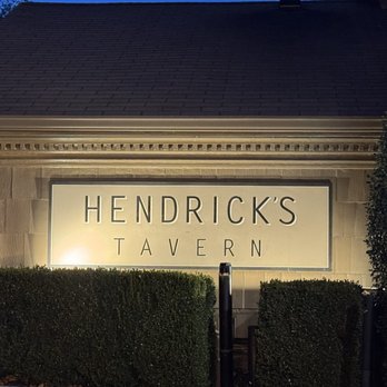 Hendrick's Tavern - The image shows an illuminated sign reading 'Hendrick's Tavern' mounted on the exterior wall of a building. The sign is backlit, making the text clearly visible against the wall. Surrounding the sign are neatly trimmed hedges, and the building has a dark, sloped roof. The scene appears to be taken during the evening or night, with the lighting emphasizing the sign and the landscaping. - Bars Near Me - Roslyn, New York