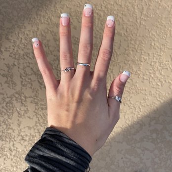 Spectrum Nails - French dip with rhinestones - A hand with manicured nails featuring white tips and small rhinestones is displayed, wearing multiple rings, with a textured beige wall in the background. - Nail Salons Near Me - Phoenix, Arizona