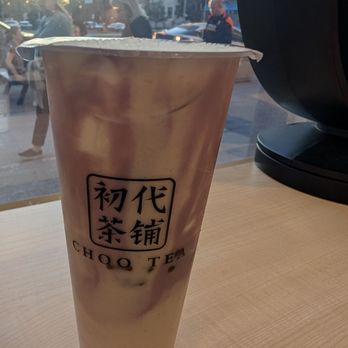 Choo Tea - Yam drink - A large paper cup containing a yam drink from Choo Tea in Toronto, ON, is positioned on a light wooden table. The cup features black Chinese characters and the words 'CHOO TEA' printed on it. The background shows a window with a view of city buildings, pedestrians walking outside, and parked cars, indicating an urban setting. The image captures the drink in a casual indoor environment with natural light coming through the window. - Bubble Tea Near Me - Toronto, Ontario