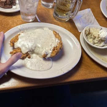 Texas Roadhouse - Country fried chicken (hand for scale), mashed potatoes with butter, green beans - A plate of country fried chicken topped with white gravy, accompanied by mashed potatoes with butter, and green beans, is placed on a wooden table. A hand with a wedding ring is visible near the plate. Additional drinks and side dishes, including a salad, are seen in the background. The setting appears to be a casual dining restaurant. - Steakhouses Near Me - St. Augustine, Florida