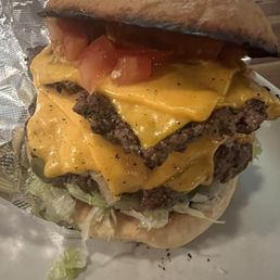 Nom nom nom - A large cheeseburger featuring multiple beef patties topped with melted cheddar cheese, slices of tomato, and shredded lettuce. The burger is assembled in a bun with a slightly toasted top, and the bottom bun is visible. The burger is partially wrapped in aluminum foil, placed on a white plate, with some lettuce peeking out from underneath.