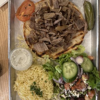 Meso Grill - Lamb gyro plate - The image shows a lamb gyro plate on a metal tray, featuring sliced cooked lamb on a piece of flatbread. Accompanying the gyro are a side salad with chopped lettuce, cucumber slices, tomato, red onion, and crumbled cheese, along with a portion of yellow rice. There is also a tomato wedge and a whole green pepper on the tray, with a small container of sauce or dressing included. The tray is placed on a wooden table. - Mediterranean Near Me - Albuquerque, New Mexico