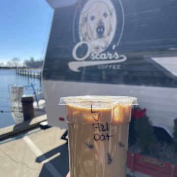 Oscar's Coffee - Iced French vanilla latte w/ oat milk. - A plastic cup of iced coffee with oat milk is held outdoors near Oscar's Coffee trailer, which features a dog logo on its side. - Coffee & Tea Near Me - Annapolis, Maryland