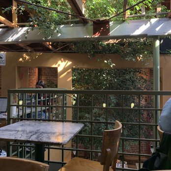 Cafe Milano - Inside - A cafe interior features a marble-topped table, wooden chairs, and a railing overlooking a lush, green outdoor space with trees and plants. - Cafes Near Me - Berkeley, California