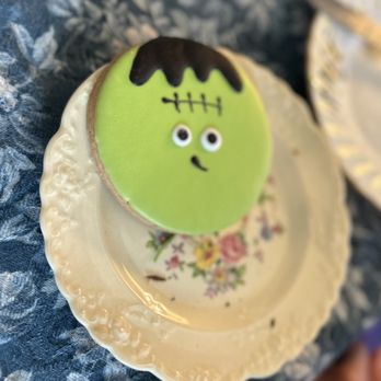 Lovey's Tea Shoppe - Halloween sugar cookie - A Halloween sugar cookie decorated with green icing to resemble Frankenstein's monster, featuring black icing for hair and facial details, with googly eyes and a small mouth. The cookie is positioned on a floral-patterned plate with a scalloped edge, set on a textured fabric surface. - British Near Me - Pacifica, California