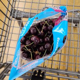 Cherries - A blue plastic bag filled with cherries is placed in a shopping cart at a grocery store, with a beige tiled floor visible.