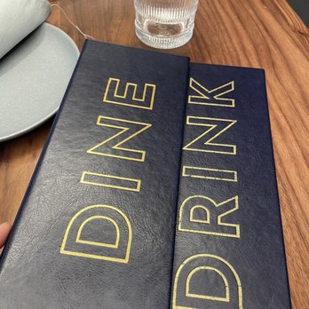 CKTL - A black, textured menu with gold outlined text reading "DINE IN" on the left and "DRINK" on the right is positioned on a wooden table. A clear, ribbed glass of water is placed behind the menu, and part of a gray plate with a napkin is visible in the upper left corner. The setting appears to be a dining or bar area. - Cocktail Bars Near Me - Toronto, Ontario