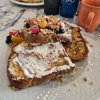 AVGO - Yogurt Fruit Parfait French Toast - A plate features two slices of French toast with a dollop of whipped cream and a topping of mixed berries, fruit, and granola. The dish is served on a white plate on a marble table, with a knife nearby. In the background, there is a plate with a dessert, a black cup, a blue AVGO branded tumbler, and an orange mug, indicating a breakfast or brunch setting at AVGO restaurant. - Breakfast & Brunch Near Me - Saint John, Indiana