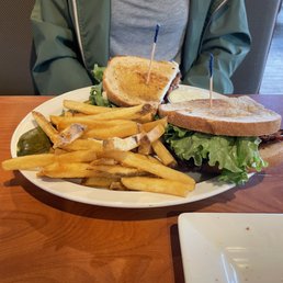 BLT Sandwich - A person in a green jacket and gray shirt is seated at a table with a large plate of sandwiches, French fries, and pickles.