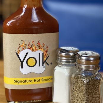 Yolk Diner - The image features a bottle of Yolk Signature Hot Sauce with a beige label and a flame design, positioned on a wooden surface. Next to the hot sauce are salt and pepper shakers made of glass with metal caps, containing salt and ground pepper respectively. The background is blurred, showing a blue object and some indistinct elements, emphasizing the condiments in the foreground. - Breakfast & Brunch Near Me - River Forest, Illinois