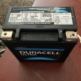 Batteries Plus - The bad battery - A Duracell Ultra platinum AGM power sport battery is shown resting on a wooden surface. The battery has warning labels indicating danger and poison, with instructions for handling and safety precautions. It features a black casing with a blue and white label displaying the Duracell logo and product details, including part number DURAGM-14 and specifications such as CCA 220. The battery terminals are visible on the top, with one terminal showing signs of corrosion or wear. - Battery Stores Near Me - Gainesville, Georgia