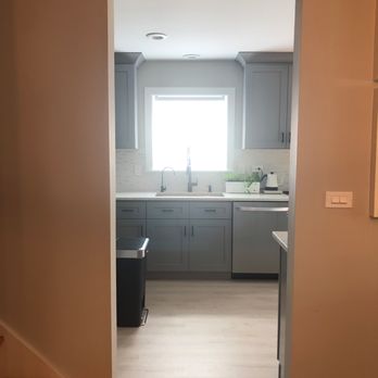 Premier Remodeling Services - A kitchen with gray cabinetry, a window, and a countertop with a plant and appliances, viewed through an open doorway. - Flooring Near Me - Morton Grove, Illinois