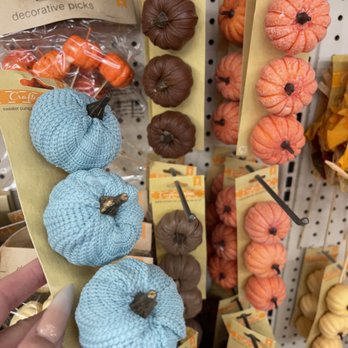 Dollar Tree - The image shows a hand holding a yellow card with three fabric pumpkins in blue, brown, and orange. These decorative pumpkins are made of textured fabric and have small brown stems. They are displayed on a pegboard with other similar decorative pumpkins in various colors, including orange and brown, arranged vertically with hooks. The setting appears to be a retail store selling decorative items, with packaging and labels visible. - Discount Store Near Me - Placentia, California