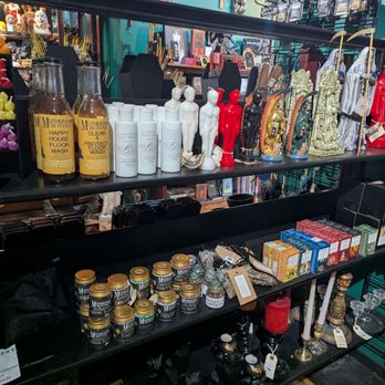 Nerdy by Nature - The image shows a multi-tiered black display shelf in a store, featuring candles, decorative figurines, small bottles, and assorted trinkets. The top shelf has bottles labeled 'Mother Botanica' and various figurines, including human-shaped and religious statues. The middle shelf contains small jars, boxes of tea, and other small items. The background reveals more store items and a colorful, busy retail environment. - Home Decor Near Me - Troy, New York