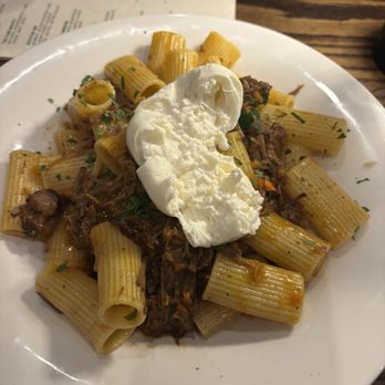 The Foundry - Short rib pasta special - A white plate contains cooked rigatoni pasta with a beef ragu sauce, garnished with chopped herbs. A poached egg is placed on top of the pasta. The dish is set on a wooden table with a menu partially visible in the background. - New American Near Me - Hicksville, New York