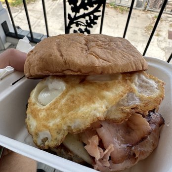 The West Egg - Cuban breakfast sandwich! - A breakfast sandwich featuring a fried egg, ham, and bread is shown in a takeout container, with an outdoor setting in the background. - Coffee & Tea Near Me - St. Augustine, Florida