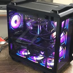 4090 i9 13900 - A custom-built gaming PC features RGB lighting on the fans, graphics card, and interior components. The case has a glass side panel, revealing the hardware inside, including a GeForce GTX graphics card and a liquid cooling system. The setup is placed on a wooden surface with a cluttered background of tools and electronic parts.