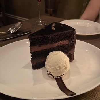 Mooo.... Beacon Hill - chocolate cake - A piece of chocolate cake with multiple layers and a glossy chocolate glaze, served with a scoop of vanilla ice cream on a white plate. - Steakhouses Near Me - Boston, Massachusetts