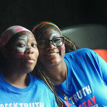 Fab Fun - Two women are close together, smiling at the camera. They are wearing blue T-shirts with the words 'Seek Truth, Stay' visible. One woman has face paint resembling a skeleton or Day of the Dead design, and the other has face paint with floral or decorative patterns. Both women are wearing glasses, and one has a headband. The background is dark, with a black surface and a red object partially visible behind them. - Game Truck Rental Near Me - Chicago , Illinois