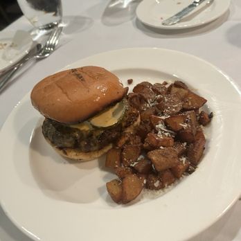 Artisan - A burger with a toasted bun, pickles, and a beef patty is placed on a white plate. Next to the burger, there are fried potato chunks sprinkled with grated cheese. The table setting includes utensils, glasses of water, and small plates, indicating a dining environment. - French Near Me - Elkhart, Indiana