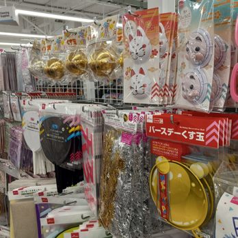 Daiso - The image shows a retail store aisle with various party supplies hanging on hooks. Items include gold-colored balloons, masks with animal and face designs, metallic streamers in gold and silver, and birthday-themed decorations. The store has a high ceiling with fluorescent lighting, and a sign indicating cleaning supplies is visible in the background. The display is organized with different types of decorations grouped together for easy browsing. - Discount Store Near Me - Aurora, Colorado