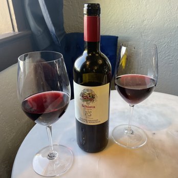 Pilon - A bottle of red wine labeled 'Schiava' is positioned on a white marble table, with two wine glasses filled with red wine on either side. The setting appears to be indoors, near a window with a blue curtain, and the background includes a textured wall and a dark blue upholstered chair. The scene suggests a casual wine-drinking moment. - Beer, Wine & Spirits Near Me - El Paso, Texas