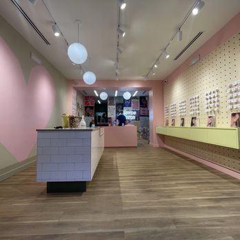 Rowan The Shoppes at Bethesda - 1.17.25 - The image shows the interior of a retail or beauty store with pastel pink and green walls. There is a white tiled counter in the foreground and display shelves on the right wall holding product images. The ceiling has track lighting and hanging spherical lights, with a visible back area that appears to be a checkout or service counter. The flooring is wood, and the space is well-lit and organized. - Jewelry Near Me - Bethesda, Maryland