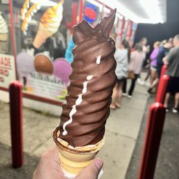 Large soft serve - A person holds a large chocolate soft serve ice cream cone with a chocolate coating and a chocolate piece on top. The cone is in front of an outdoor ice cream stand with a line of people waiting. The stand has a sign with images of ice cream cones and a neon sign. The scene is illuminated by bright lighting, and the background shows a crowd of people in casual clothing.