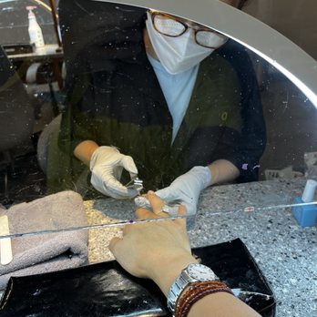 Nails Uncorked Amazon - A client is getting a manicure at a nail salon, with the technician working through a glass partition, wearing gloves and a mask. - Nail Salons Near Me - Eugene, Oregon