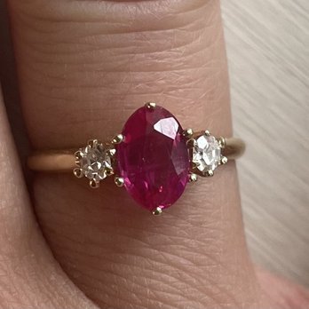 Mark's Diamonds - A gold ring features a central oval ruby flanked by two small diamonds, set on a person's finger. - Jewelry Near Me - Denver, Colorado