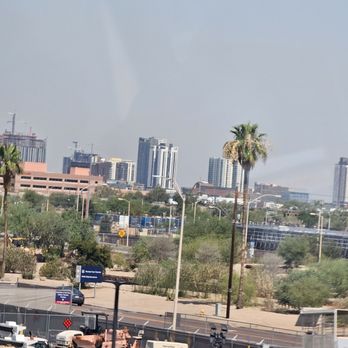 PHX Sky Train - View of Phoenix on the train. - The image shows a cityscape of Phoenix, Arizona, as seen from the PHX Sky Train. Tall buildings and skyscrapers are visible in the background, with some construction cranes present. Palm trees are in the foreground, along with streetlights, parking areas, and a fence. The sky is partly cloudy, and the overall scene captures an urban environment with natural desert elements. - Trains Near Me - Phoenix, Arizona