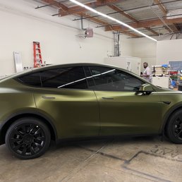 Avery Hope Green satin wrap - A Tesla Model Y features a matte green vinyl wrap, with tinted windows and black wheels, parked inside a workshop or industrial space. The background includes workbenches, tools, and a person wearing a mask, indicating an environment for vehicle customization or wrapping services.