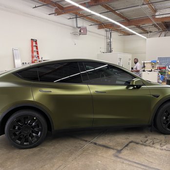 Big Boy Wraps - Avery Hope Green satin wrap - A Tesla Model Y features a matte green vinyl wrap, with tinted windows and black wheels, parked inside a workshop or industrial space. The background includes workbenches, tools, and a person wearing a mask, indicating an environment for vehicle customization or wrapping services. - Vehicle Wraps Near Me - Martinez, California