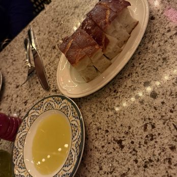 Valentina's - Bread - The image shows a white oval plate with several slices of bread, which appear to be toasted or baked, placed on a speckled granite countertop. Next to the plate is a decorative bowl containing a yellow liquid, possibly olive oil or a similar condiment. The setting suggests a dining environment, with utensils and a chair visible in the background. - Italian Near Me - Dublin, Ohio
