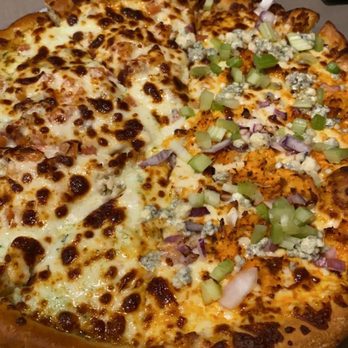 Mountain Mike's Pizza - A large pizza features melted cheese and grilled chicken on one half, with a golden-brown crust. The other half is topped with chopped onions, green peppers, and crumbled blue cheese, creating a two-tone appearance. The pizza is placed on a table with a dark background, and part of a green and white box is visible in the corner. - Pizza Near Me - Orem, Utah