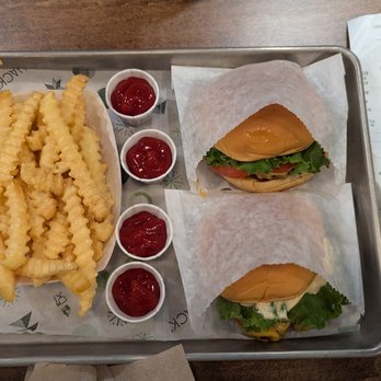 Shake Shack - A metal tray contains two sandwiches wrapped in paper, each with visible lettuce and tomato, placed on a wooden table. To the left of the sandwiches are crinkle-cut fries, and four small cups of ketchup are arranged vertically beside the fries. There are receipts and a water bottle on the table, indicating a casual dining setting. - Hot Dogs Near Me - Toronto, Ontario