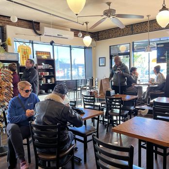 Yorgo's Bageldashery - Dining on the first floor - A bagel shop interior with customers seated at tables, some looking at menus or using phones, and a display shelf with baked goods and merchandise. - Bagels Near Me - Norfolk, Virginia