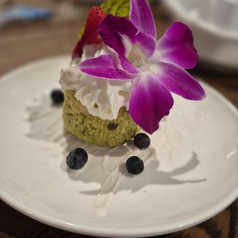 Harrison’s - Pistachio ice cream - A dessert on a white plate includes a green cake with a crumbly texture, topped with whipped cream, a strawberry, and a purple orchid flower. Several blueberries are arranged around the cake, and the plate has a drizzle of white sauce. The background is blurred, focusing on the dessert. - Steakhouses Near Me - Indianapolis, Indiana
