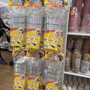 Daiso - The image shows a store display rack holding multiple plastic musubi makers in yellow packaging, each with instructions and images. The rack is situated in a retail environment with other kitchen and food containers visible on nearby shelves. The store appears to be a variety or discount store, with shoppers and shopping carts in the background. - Department Stores Near Me - Olathe, Kansas