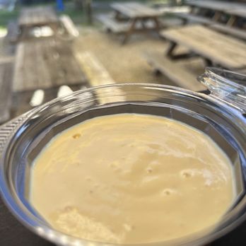 Casafinca - A clear plastic container filled with a creamy, yellowish liquid, set outdoors with wooden picnic tables and benches in the background. - Comfort Food Near Me - Miami, Florida