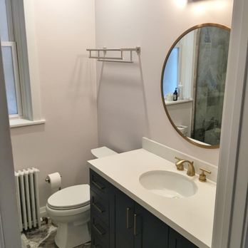 Demo Workers - A bathroom features a white vanity with gold fixtures, a round mirror, a toilet, and a window with a radiator below. - General Contractors Near Me - Hubbard, Illinois