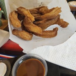 Cajeta - Crispy, fried churros served in a paper tray with a cup of cajeta sauce, typical of a Mexican-style churro shop.