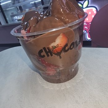 Chocolate Bash - Dubai Chocolate Strawberries Cup - A transparent plastic cup contains strawberries dipped in chocolate, with some strawberries visible at the bottom. The cup has a logo with the word 'CHOCOLAT' and a small strawberry illustration. The background shows a counter and a neon sign with a strawberry graphic, indicating a dessert or confectionery setting. - Desserts Near Me - Manteca, California